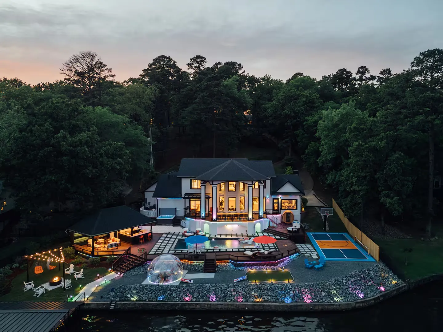 Lakefront home with a pool, sport court and string lights at dusk
